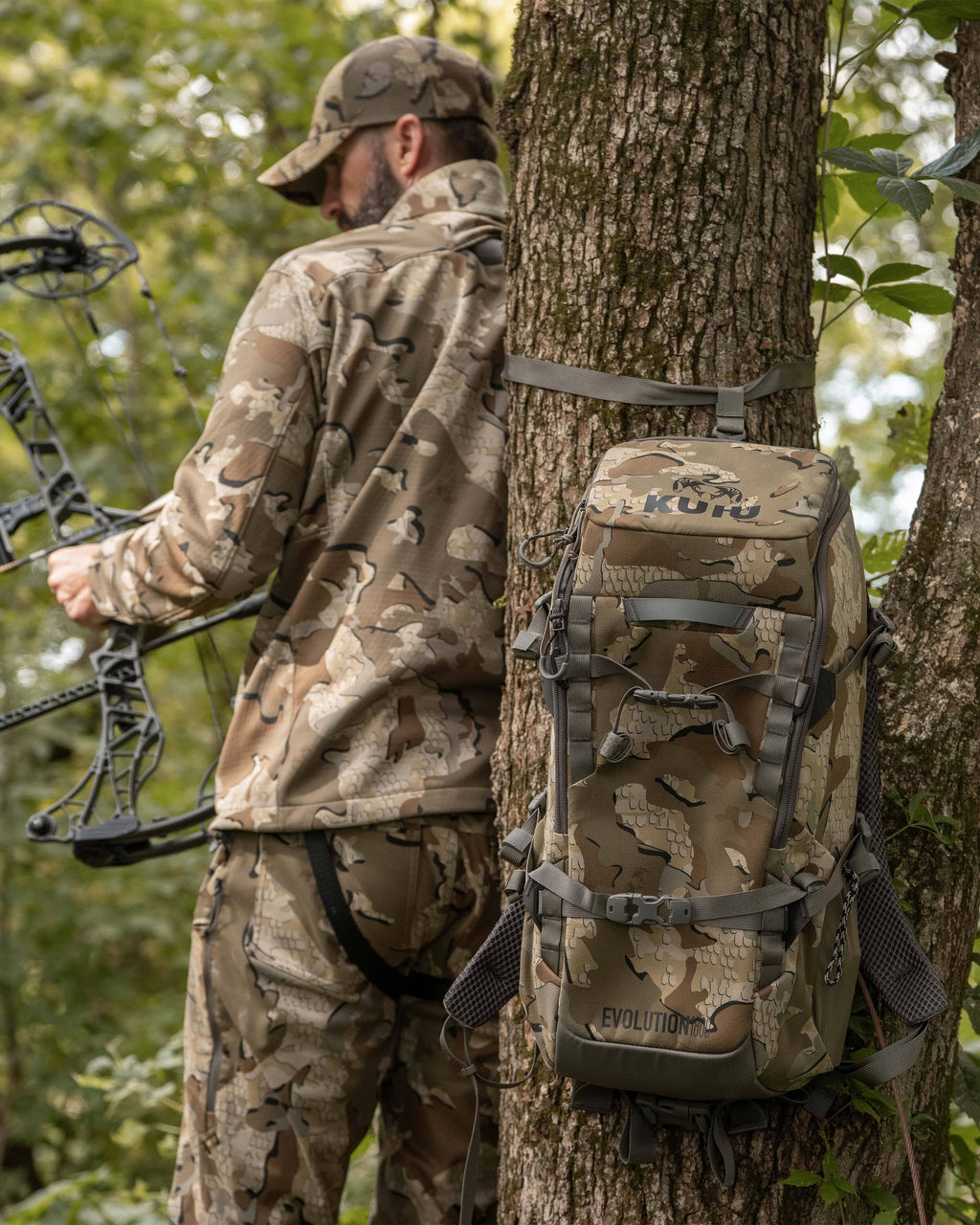 Whitetail hunter in a tree stand, stands with his back to the camera holding a compound bow in the left hand with Evolution 1800 in Valo Camouflage strapped to a tree trunk beside him