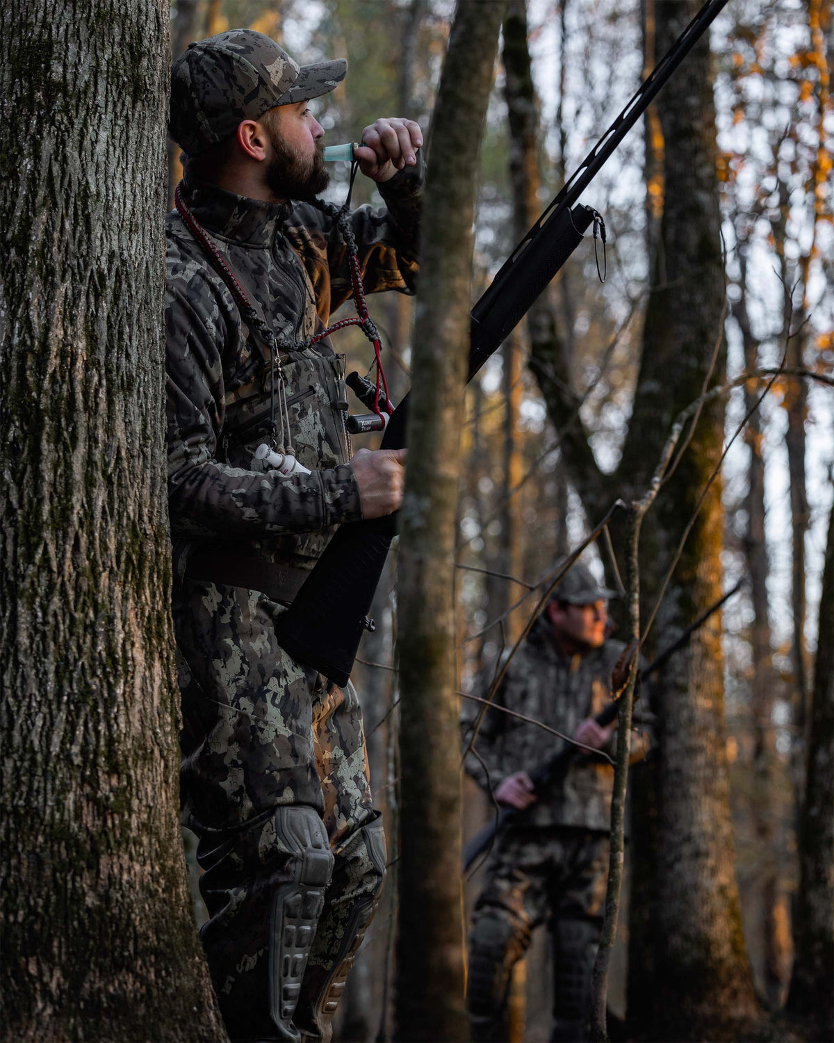 Hunter wearing StrongFleece 290 Full Zip in Veros camouflage stands behind a tree in a wooded forest, holding a bow. Another person in similar camouflage is visible in the blurred background among the trees.
