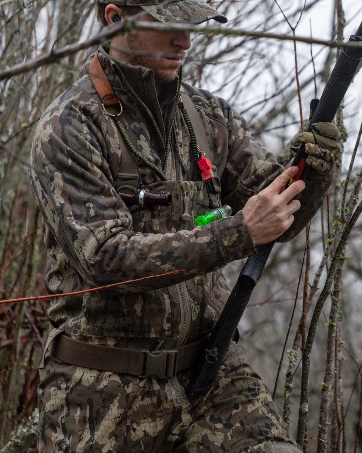 Hunter wearing StrongFleece 290 Full Zip in Veros camouflage and holds a rifle while standing among bare trees and branches outdoors.
