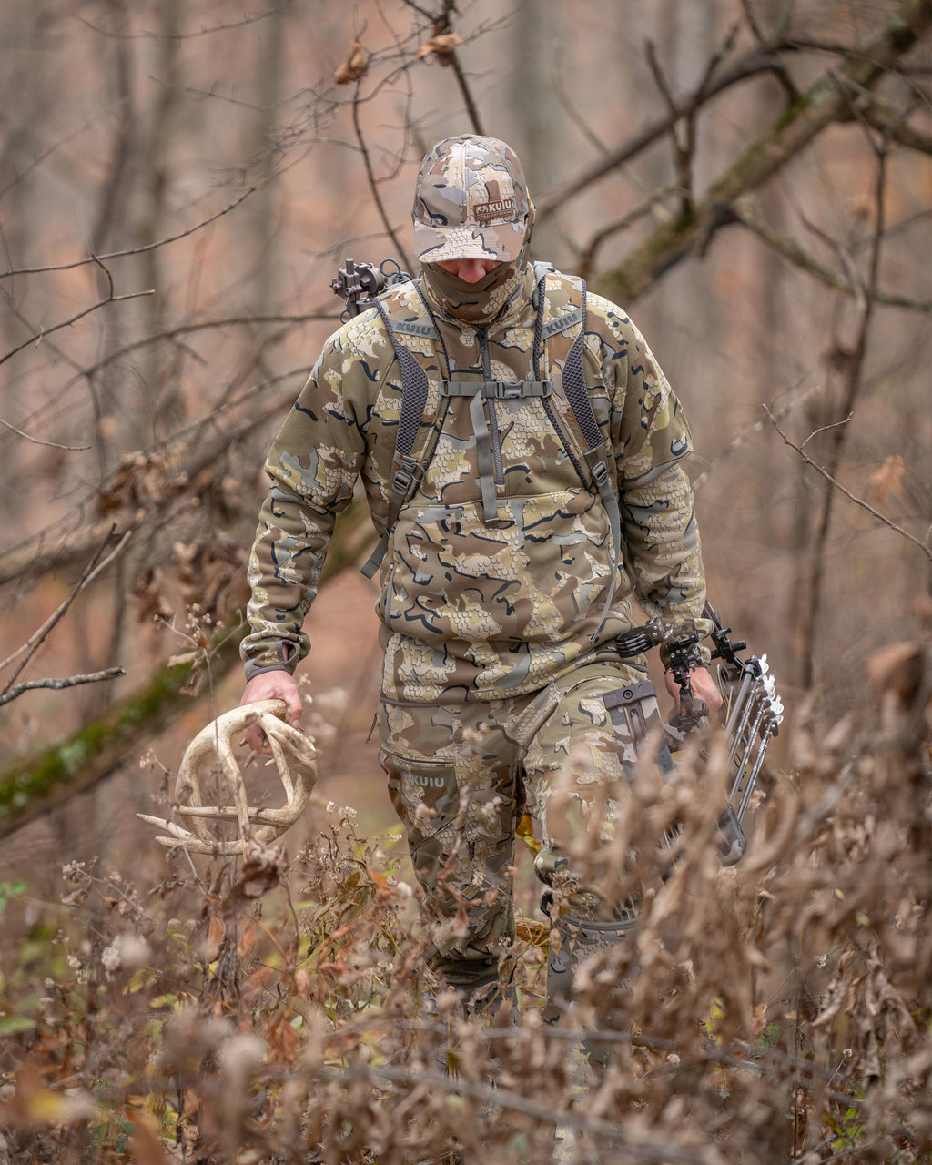 Hunter wearing Encounter 290 Fleece Zip-T in Valo Camouflage, carrying deer antlers and a bow, walking through the woods