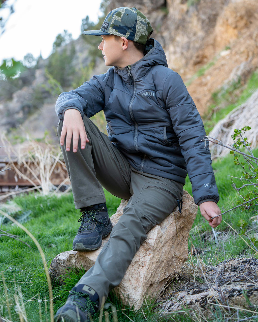 Youth wearing the Gunmetal gray side of the Youth Reversible Versa Jacket in Verde Camouflage, casually sitting on a rock looking away into the distance
