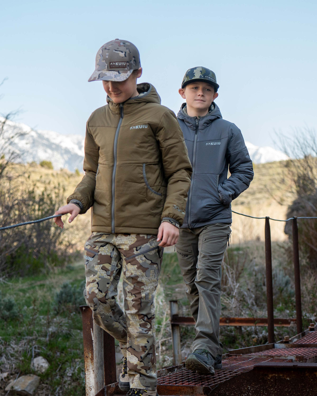 Youth boys both wearing reversible solid side of Youth Reversible Versa Jacket in Verde and Valo Camouflage, walking on a rustic bridge pathway
