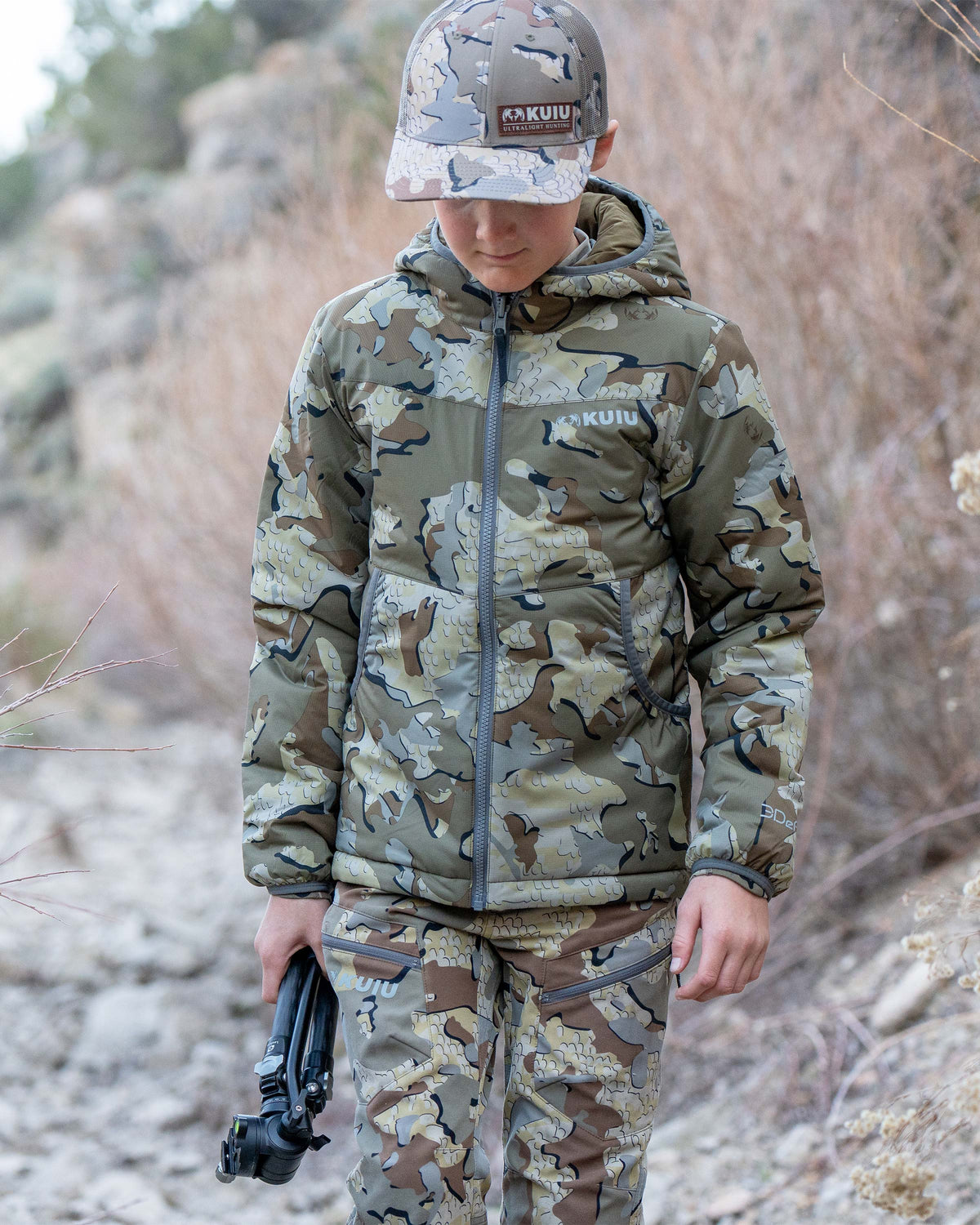 Boy wearing Youth Reversible Versa Jacket in Valo Camouflage, carrying a tripod in his right hand