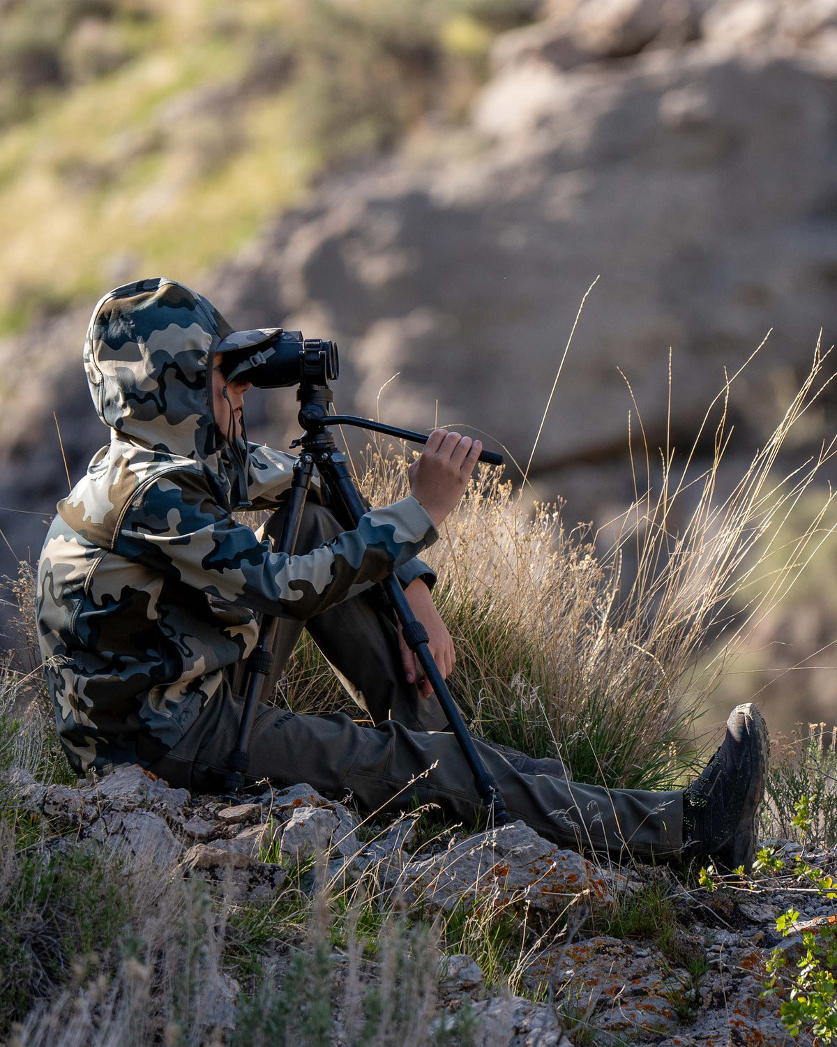 Youth sitting on a rocky terrain, using binoculars on a tripod, wearing Youth Trailblazer Jacket in Vias Camouflage
