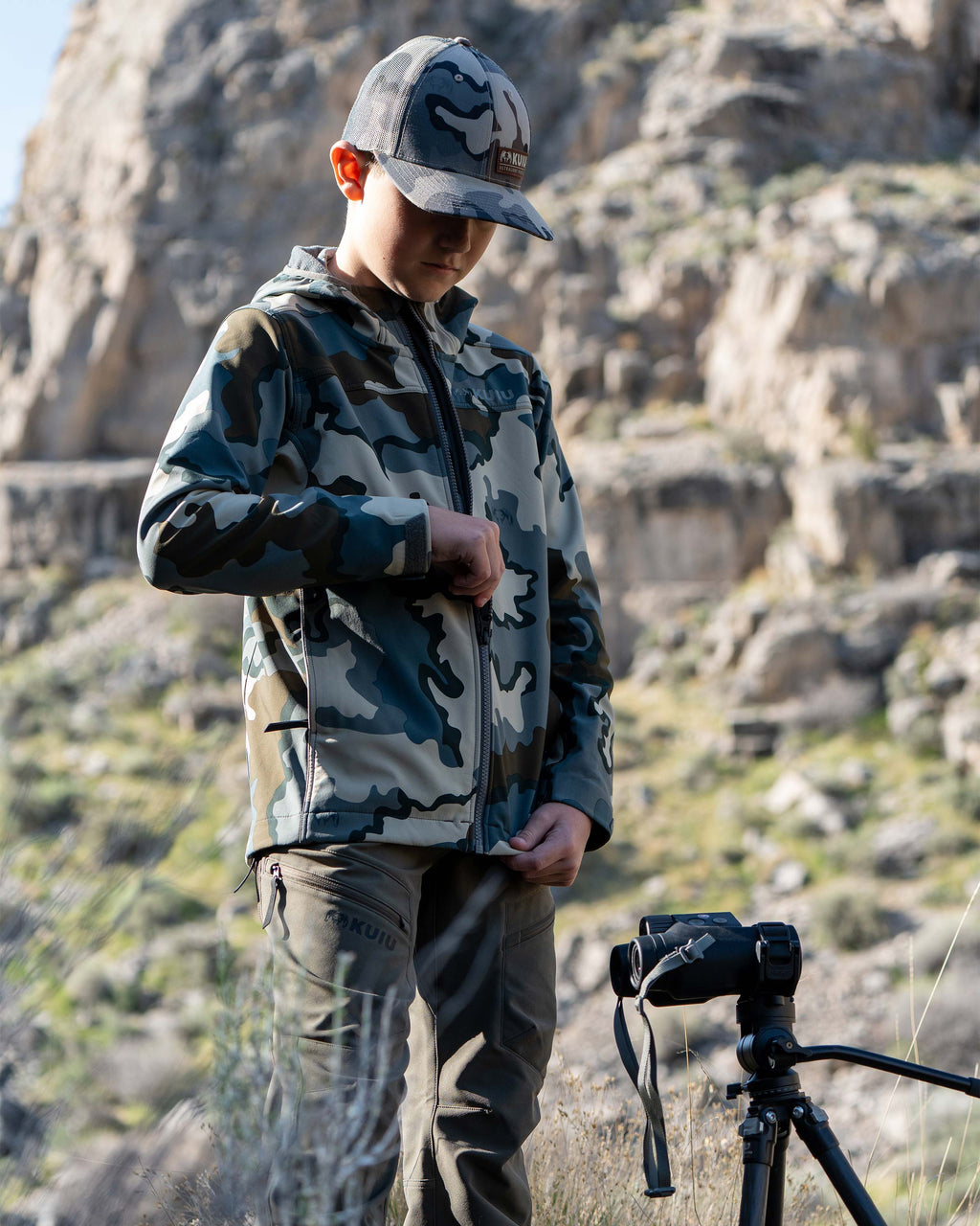 Youth zipping up his Trailblazer Jacket in Vias Camouflage, surrounded by a rocky terrain background