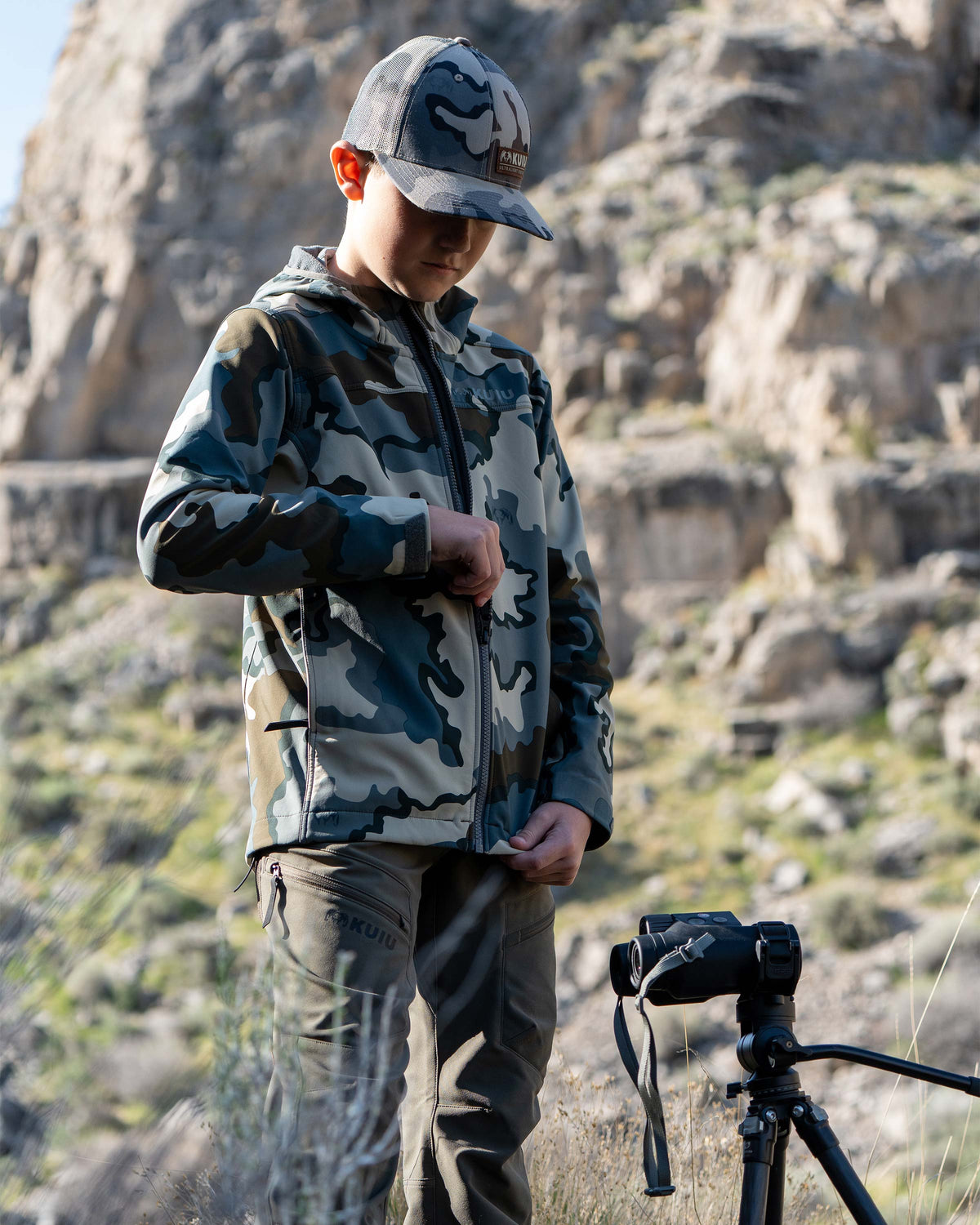 Youth zipping up his Trailblazer Jacket in Vias Camouflage, surrounded by a rocky terrain background