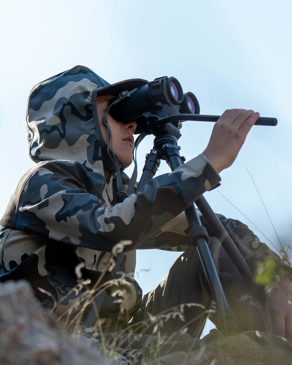 Close-up of Youth looking through his binocular on a tripod, wearing Youth Trailblazer Jacket in Vias Camouflage