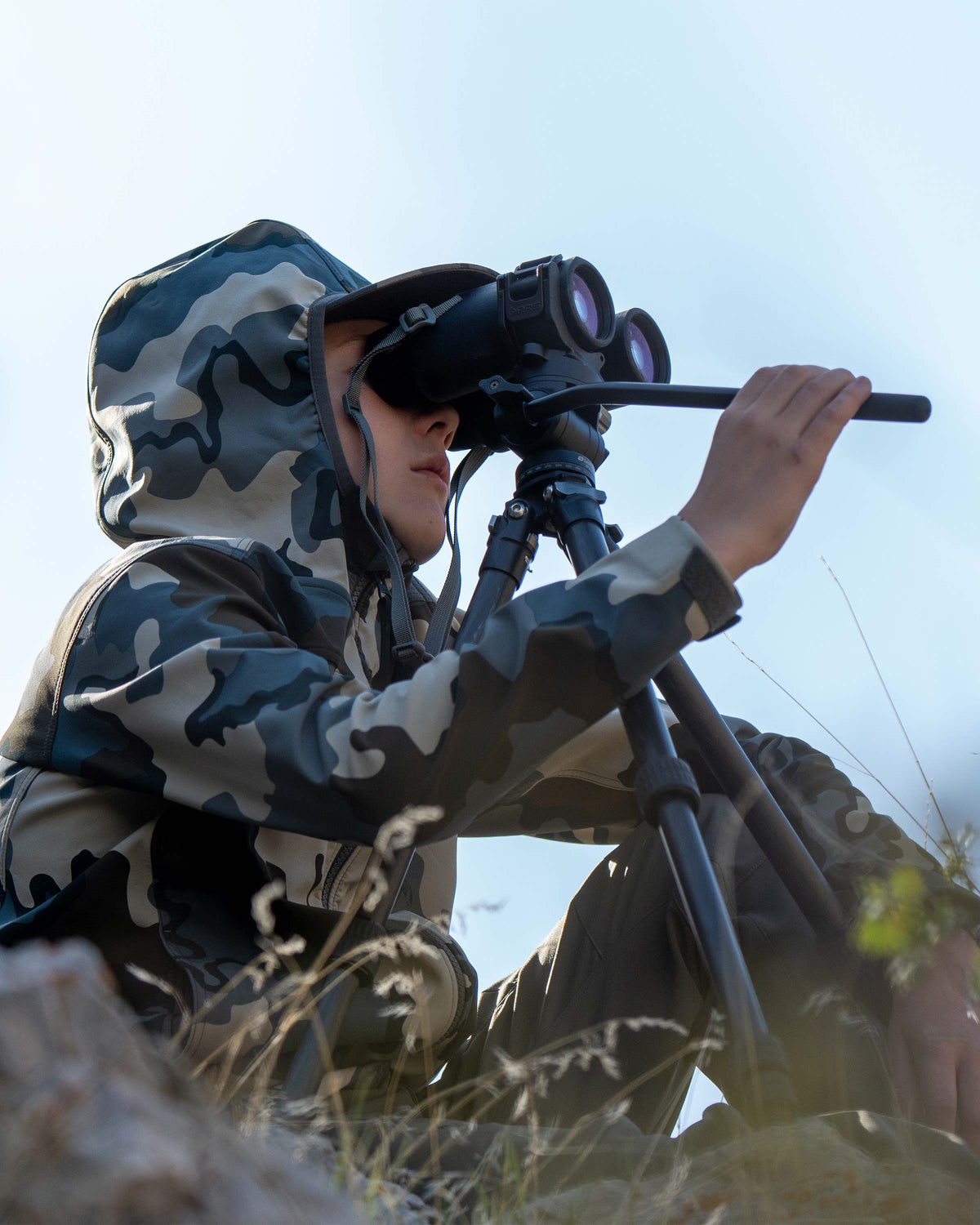 Close-up of Youth looking through his binocular on a tripod, wearing Youth Trailblazer Jacket in Vias Camouflage