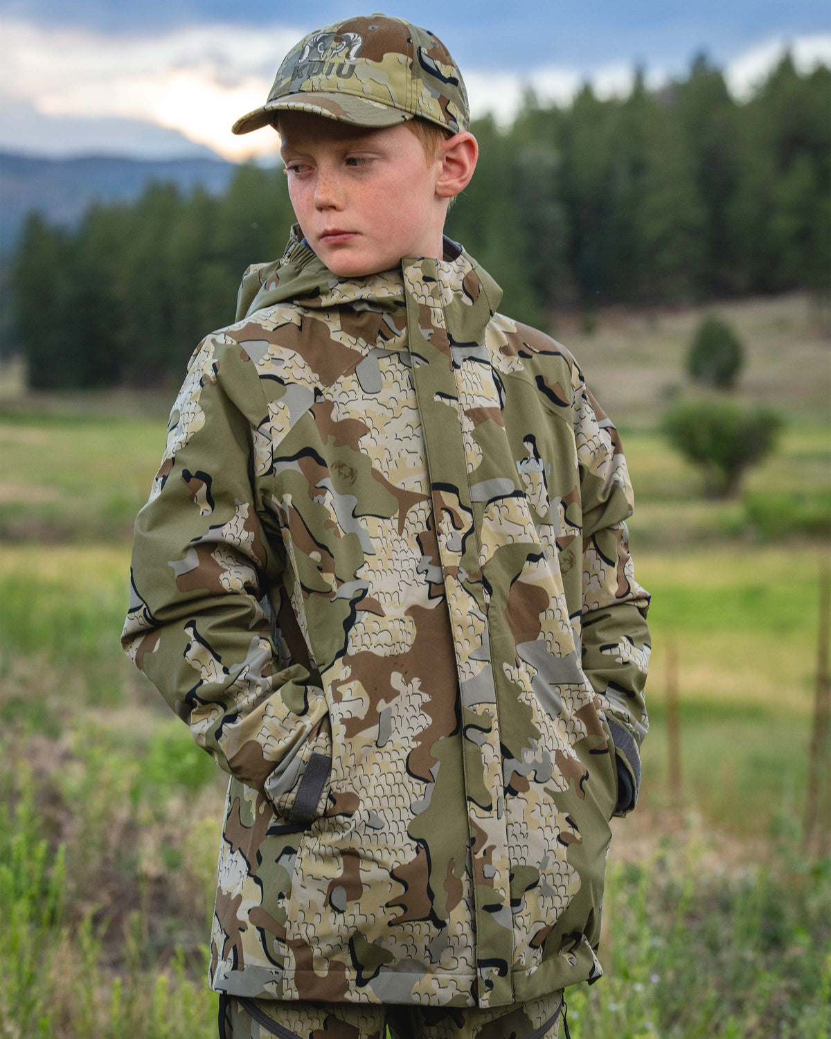 A young male teenager wearing a Youth Versa Jacket in Valo camouflage hunting jacket and matching camouflage cap, standing outdoors in a grassy field with trees in the background.