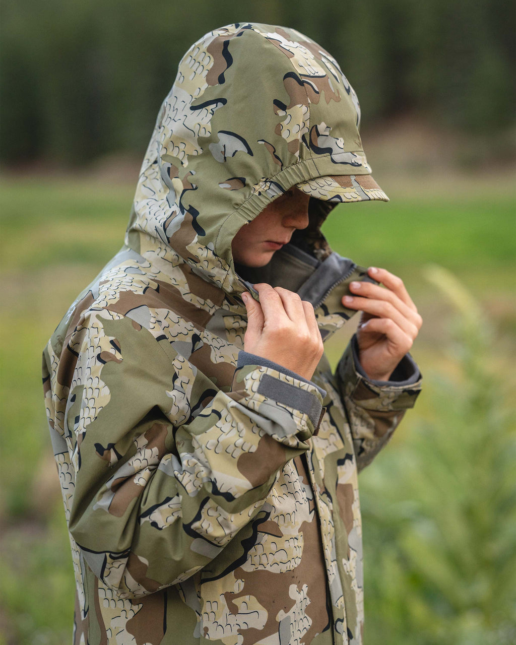 A young male teenager wearing a hooded Youth Versa Jacket in Valo camouflage outdoors, adjusting the collar with both hands in a grassy field.