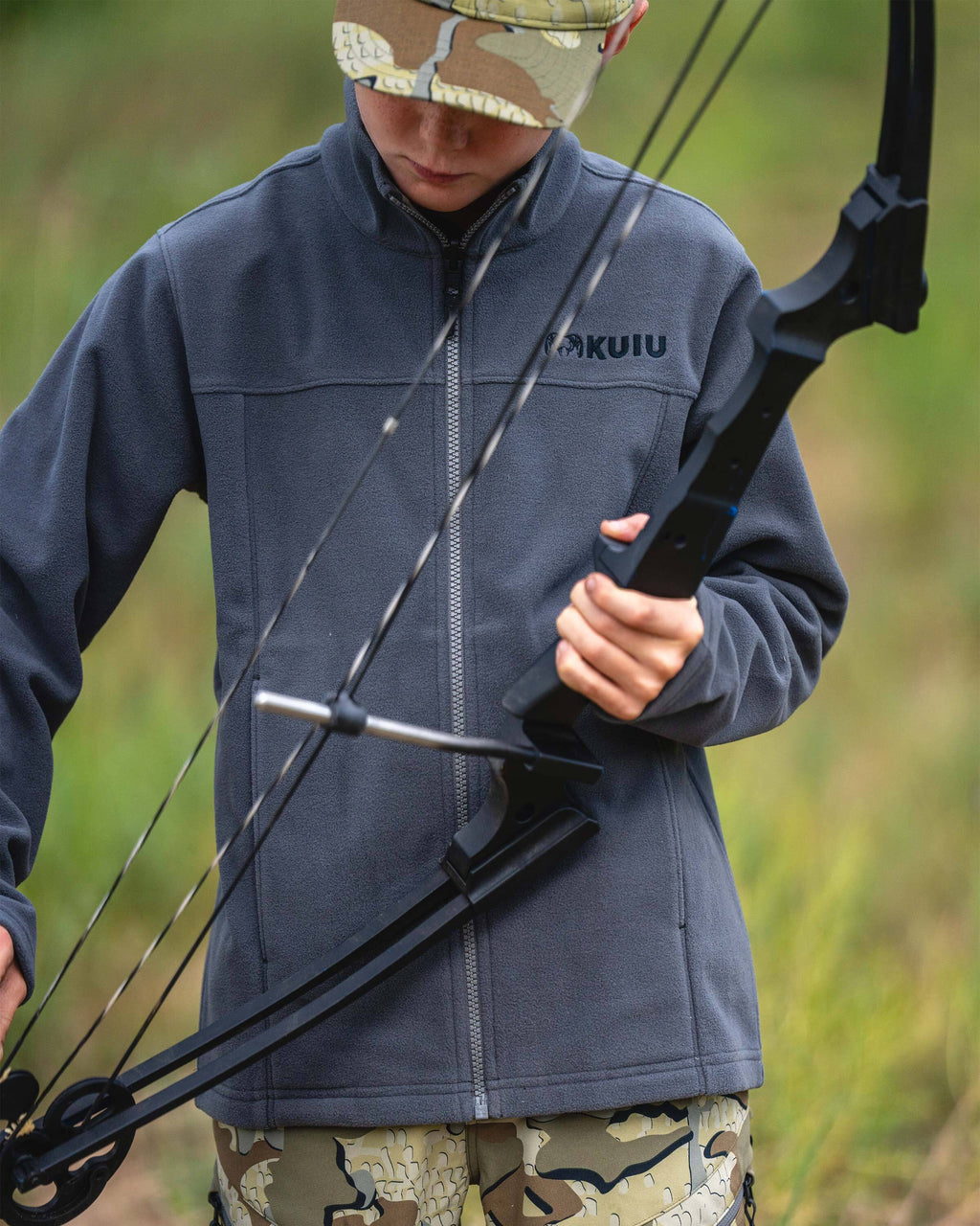 Youth wearing a Youth Versa Fleece Full Zip in Gunmetal Grey, holding a compound bow. The background is a soft-focus grassy field.