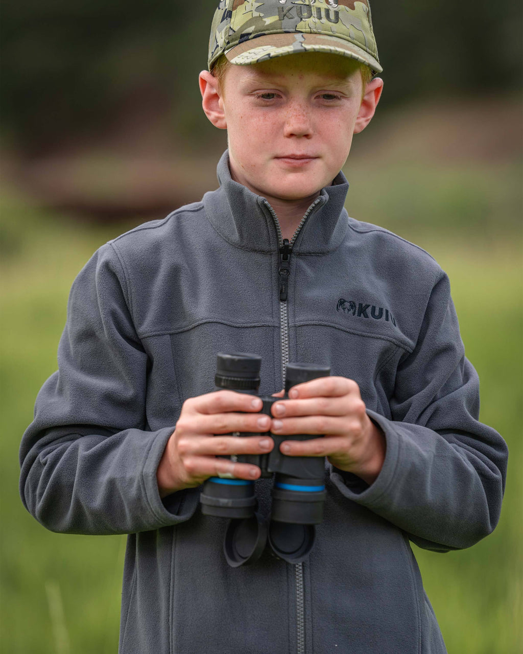 Close-up of a youth wearing a Youth Versa Fleece Full Zip in Gunmetal Grey, holding a pair of binoculars in a grassy outdoor setting.