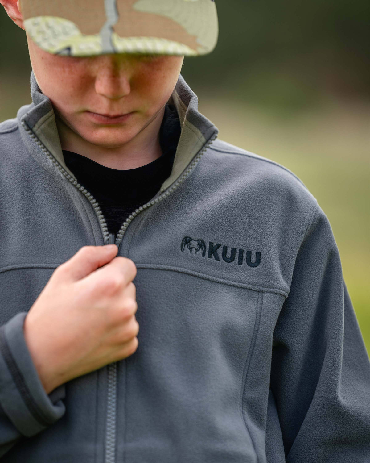Close-up of a youth wearing a Youth Versa Fleece Full Zip in Gunmetal Grey, pulling the zipper, highlighting the zipped raised collar.
