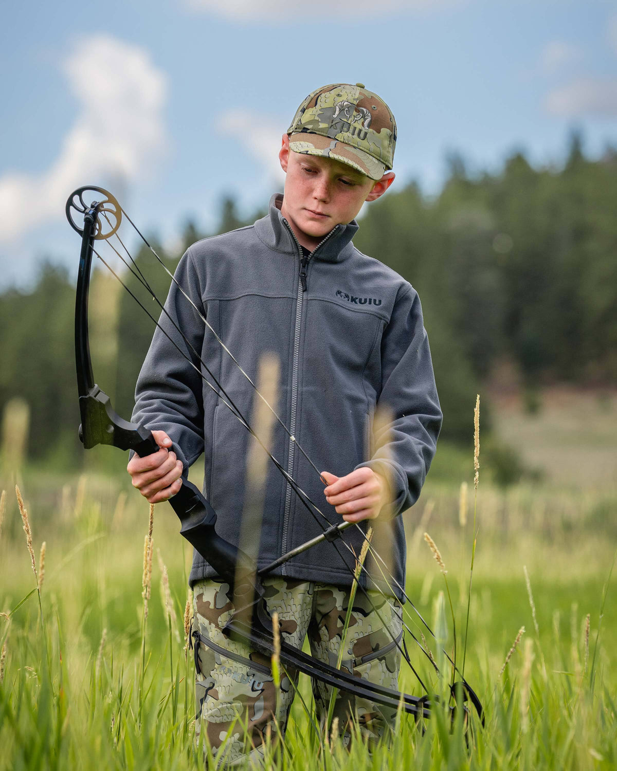 Youth stands in a grassy field holding a black compound bow, wearing a Youth Versa Fleece Full Zip in Gunmetal Grey.