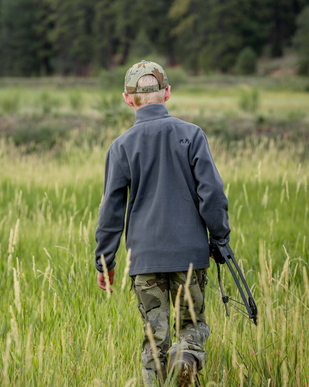 Back view of youth wearing Youth Versa Fleece Full Zip in Gunmetal Grey, walking through a tall green grassy field, holding a compound bow.