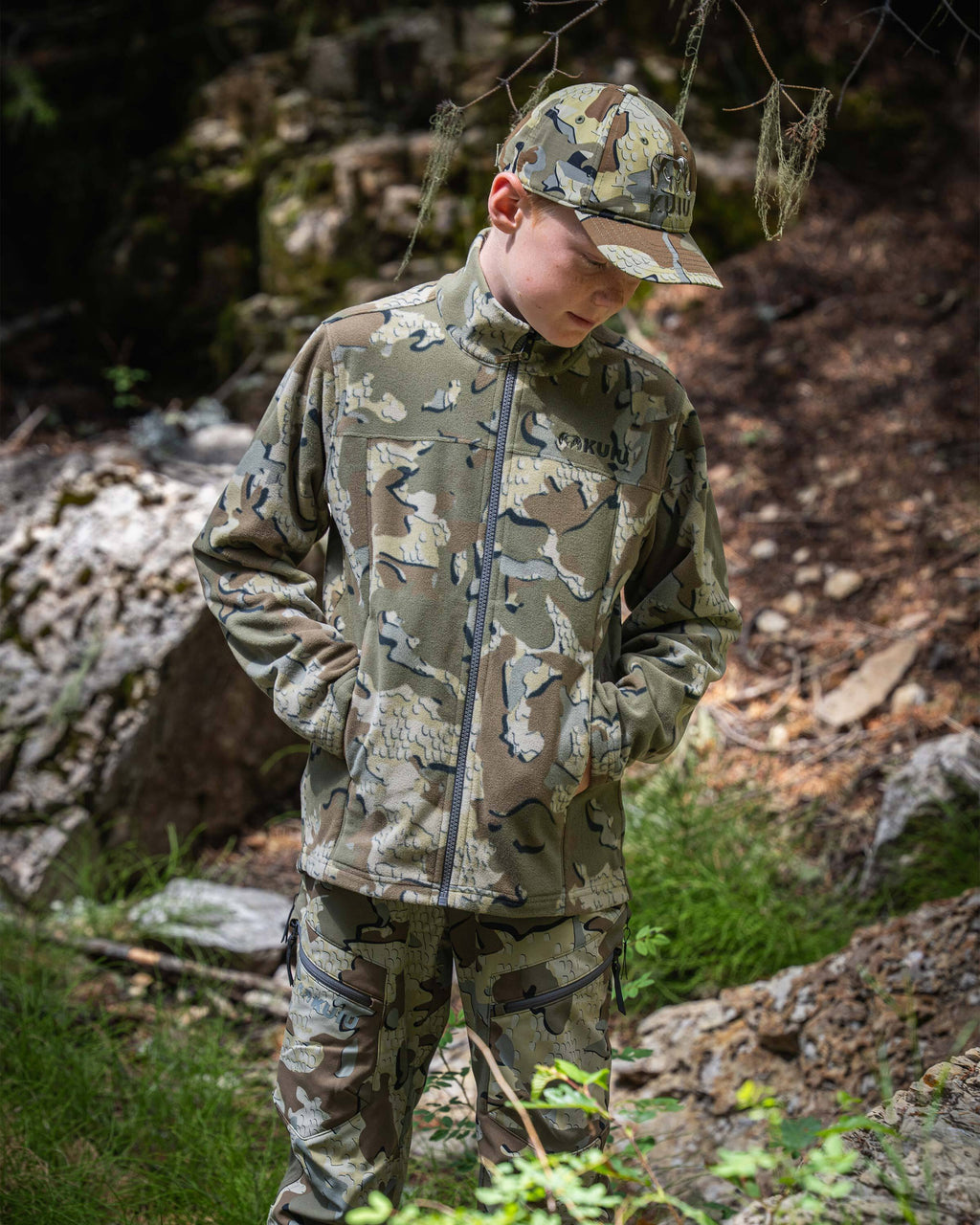 Youth hunter wearing Youth Versa Fleece Full Zip in Valo Camouflage standing in a rocky forest setting.