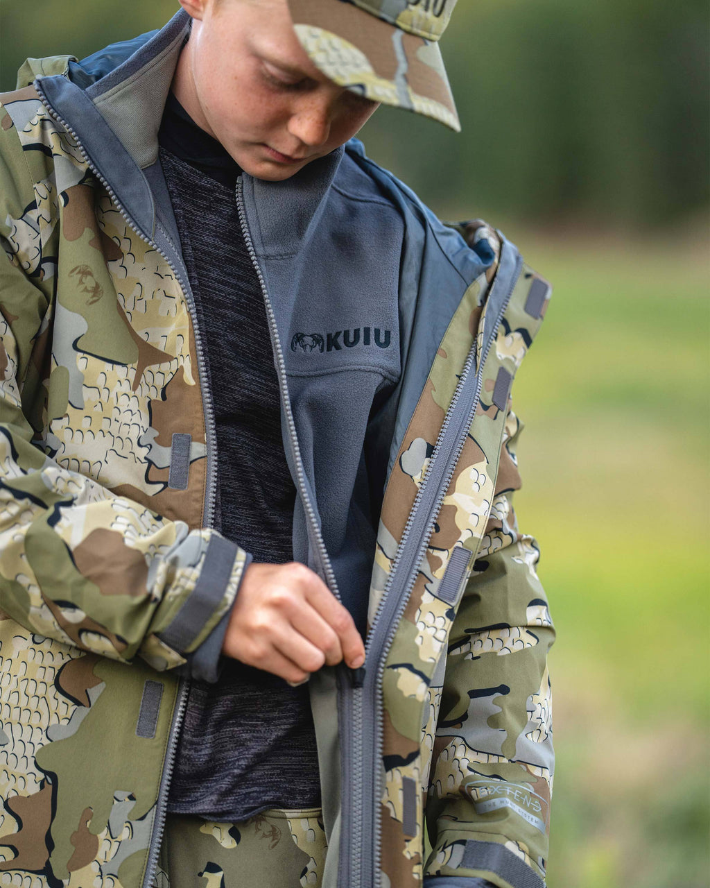 Youth hunter wearing Youth Versa Fleece Full Zip in Gunmetal Grey unzipping Youth Versa Rain Jacket, outershell liner, in Valo Camouflage.