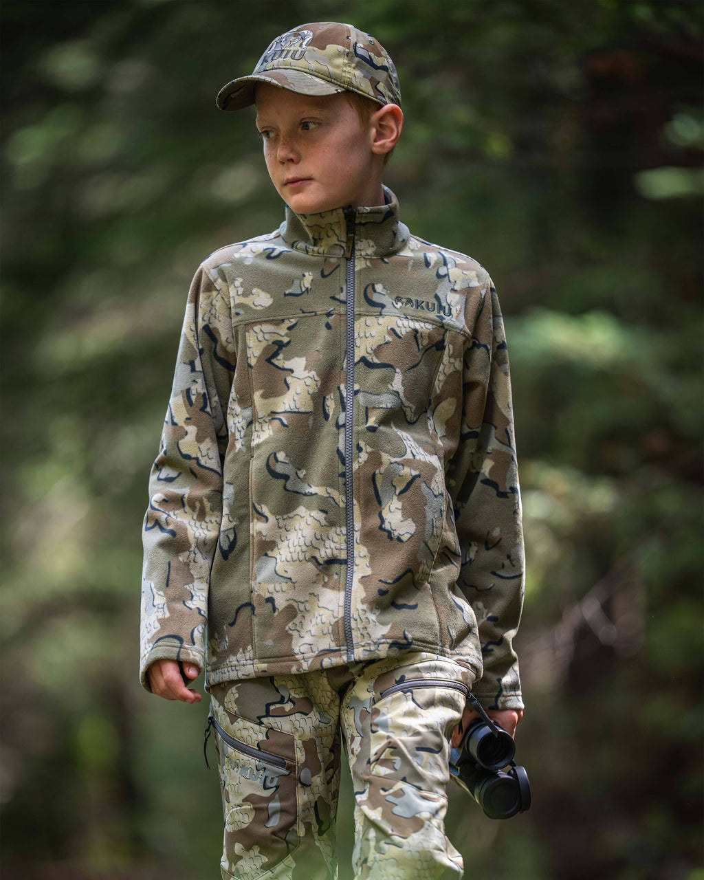 Youth hunter wearing a Youth Versa Fleece Full Zip in Valo Camouflage, standing outdoors in a forest setting and holding binoculars.