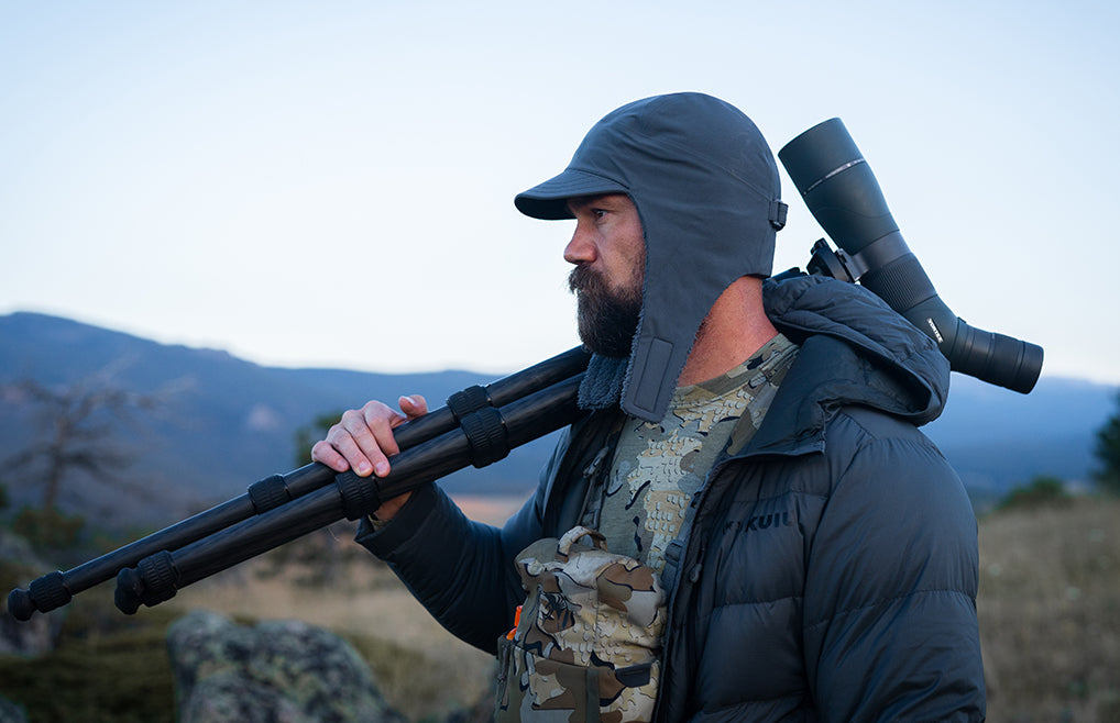 A man wearing a Mountain PRO Hat in Gunmetal Grey, carrying a black tripod over his shoulder and wearing a spotting scope on his back. 