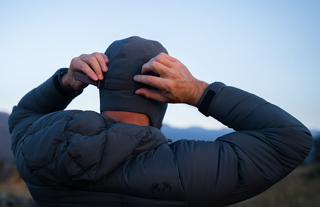 Back view of a man wearing a Mountain PRO Hat in Gunmetal Grey adjusting it with both hands, in an outdoor soft-focus landscape and a pale blue sky.