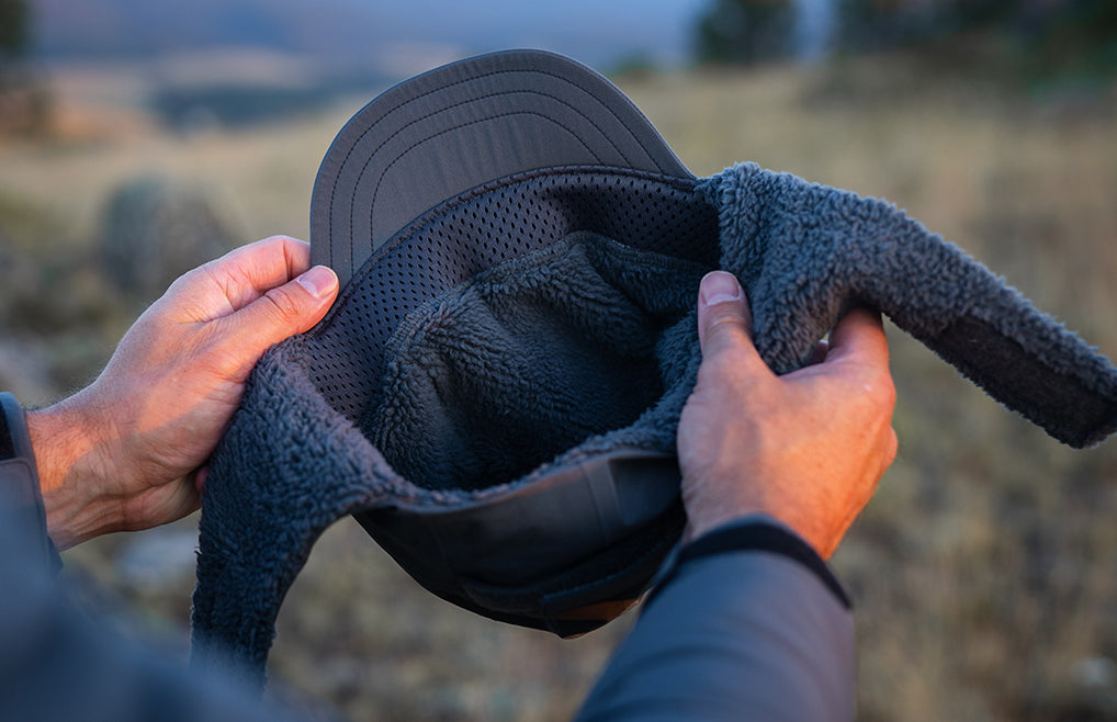 A person holds a Mountain PRO Hat in Gunmetal Grey, showing interior fleece-lined cap in an outdoor field at sunset.