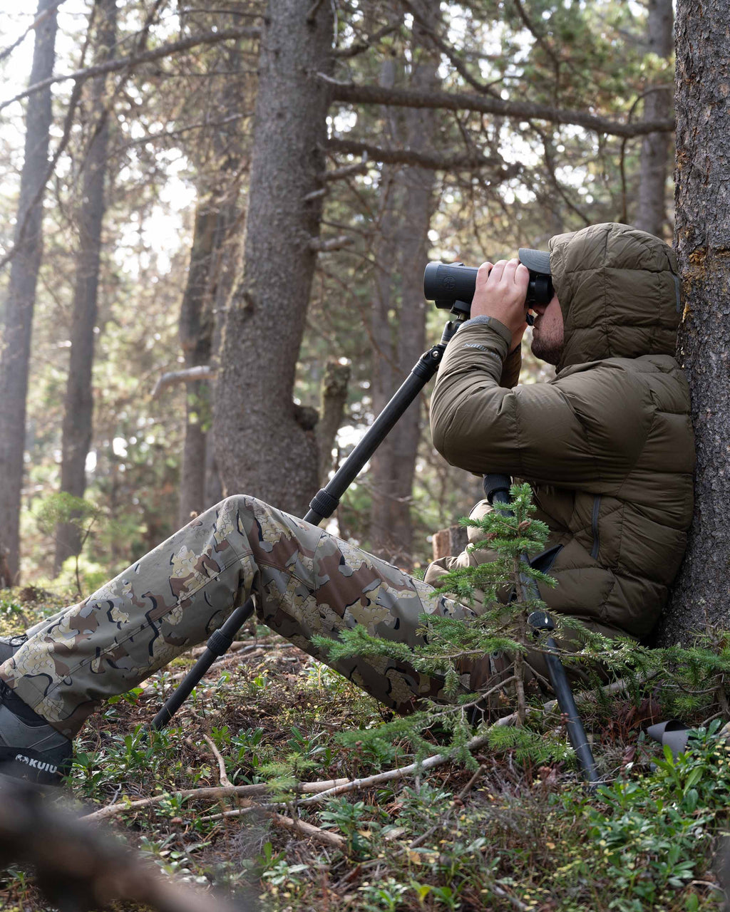 Hunter sitting and using binoculars wearing Valo Camouflage pants and the Super Down PRO Hooded Jacket in Bourbon