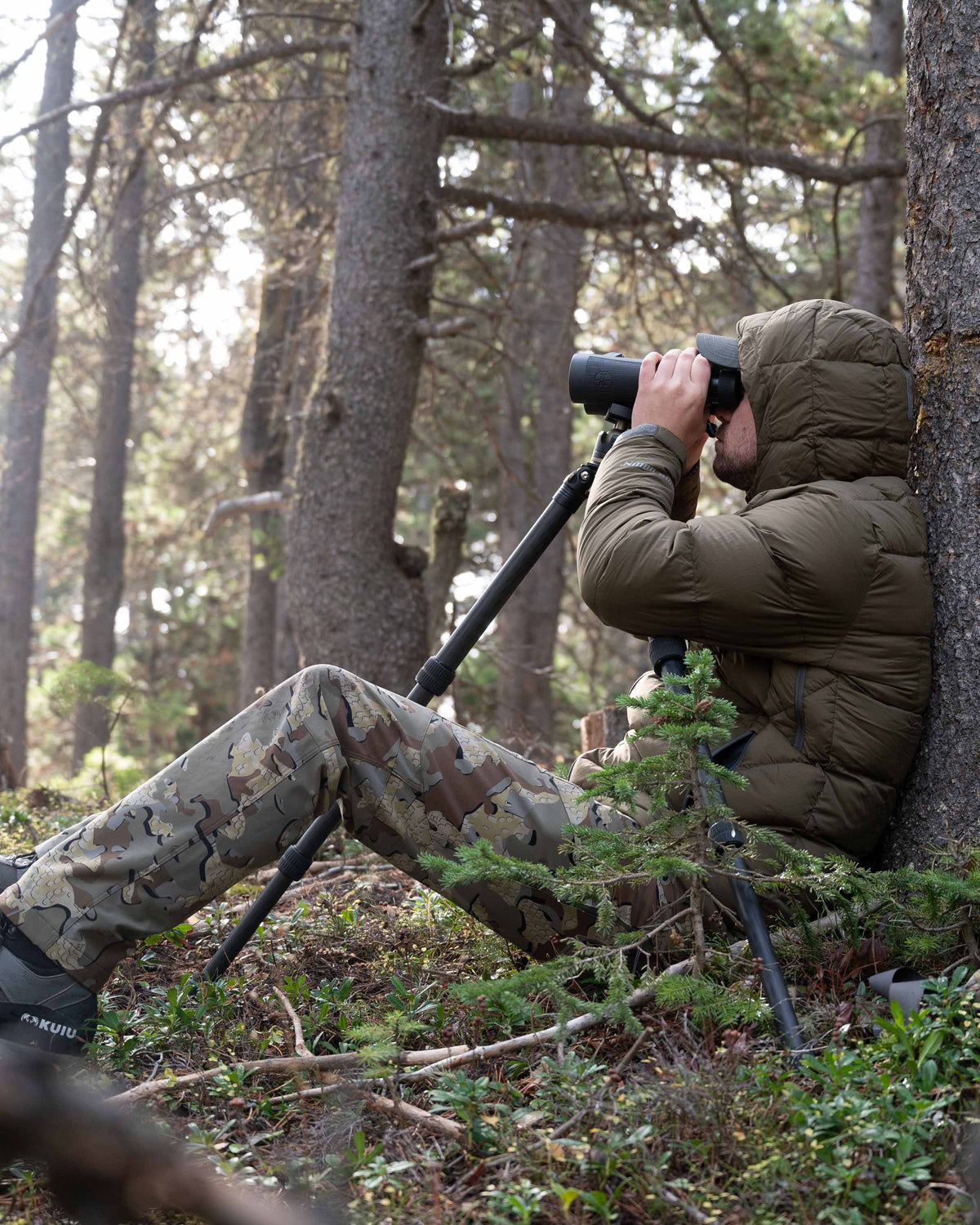 Hunter sitting and using binoculars wearing Valo Camouflage pants and the Super Down PRO Hooded Jacket in Bourbon