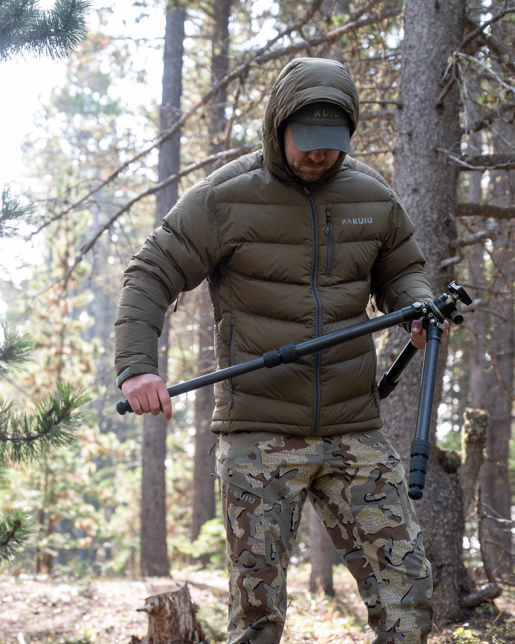 Hunter extending a tripod wearing Valo Camouflage pants and the Super Down PRO Hooded Jacket in Bourbon