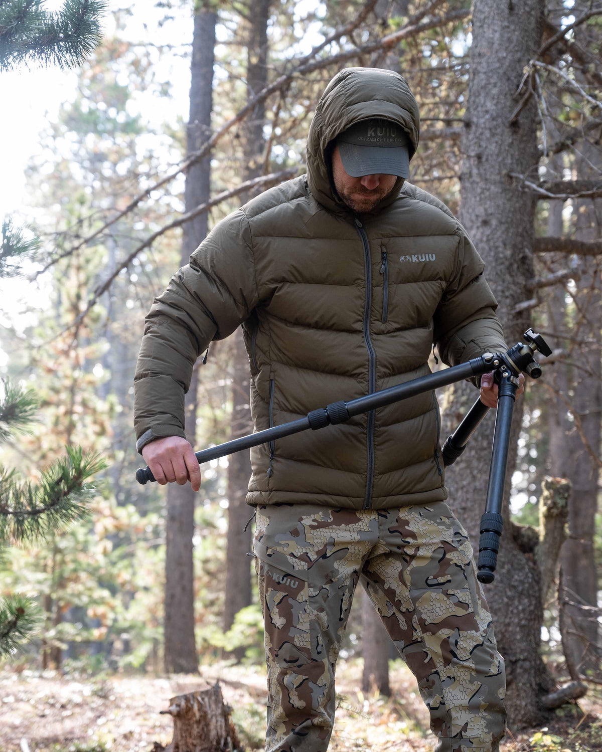 Hunter extending a tripod wearing Valo Camouflage pants and the Super Down PRO Hooded Jacket in Bourbon