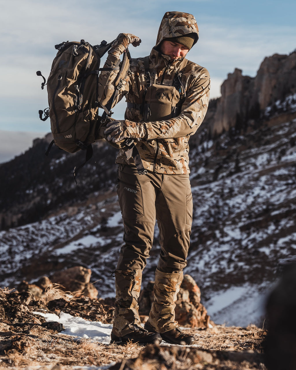 Hunter putting on a backpack while wearing the Axis Hybrid Hooded Jacket in Valo Camouflage