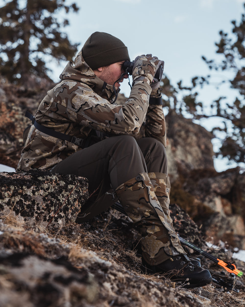 Hunter sitting and using binoculars while wearing the Axis Hybrid Hooded Jacket in Valo Camouflage