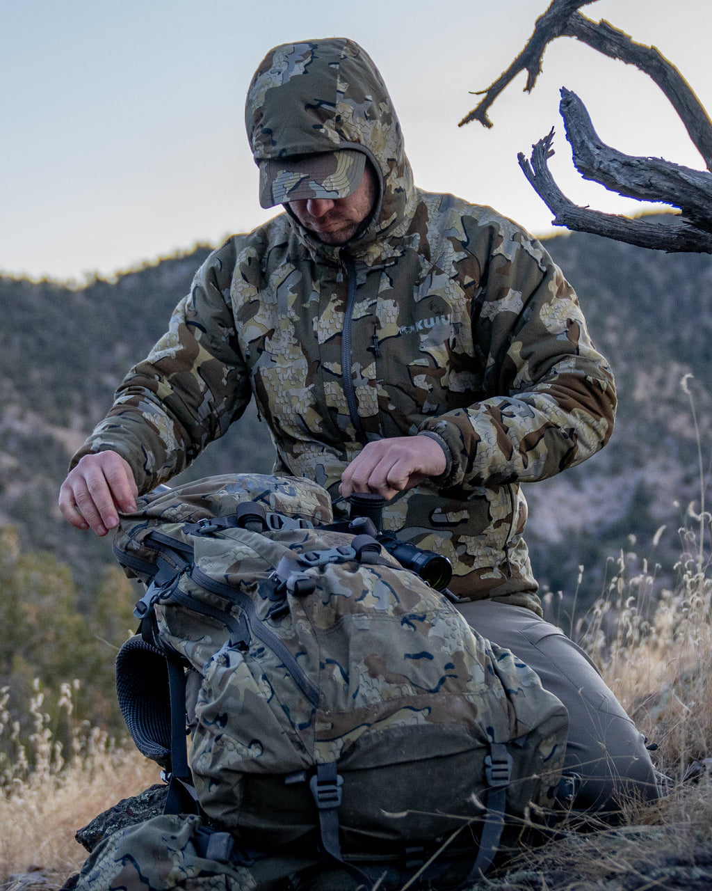 Hunter in field on knees getting into pack wearing Kenai hooded Jacket in Valo Camouflage