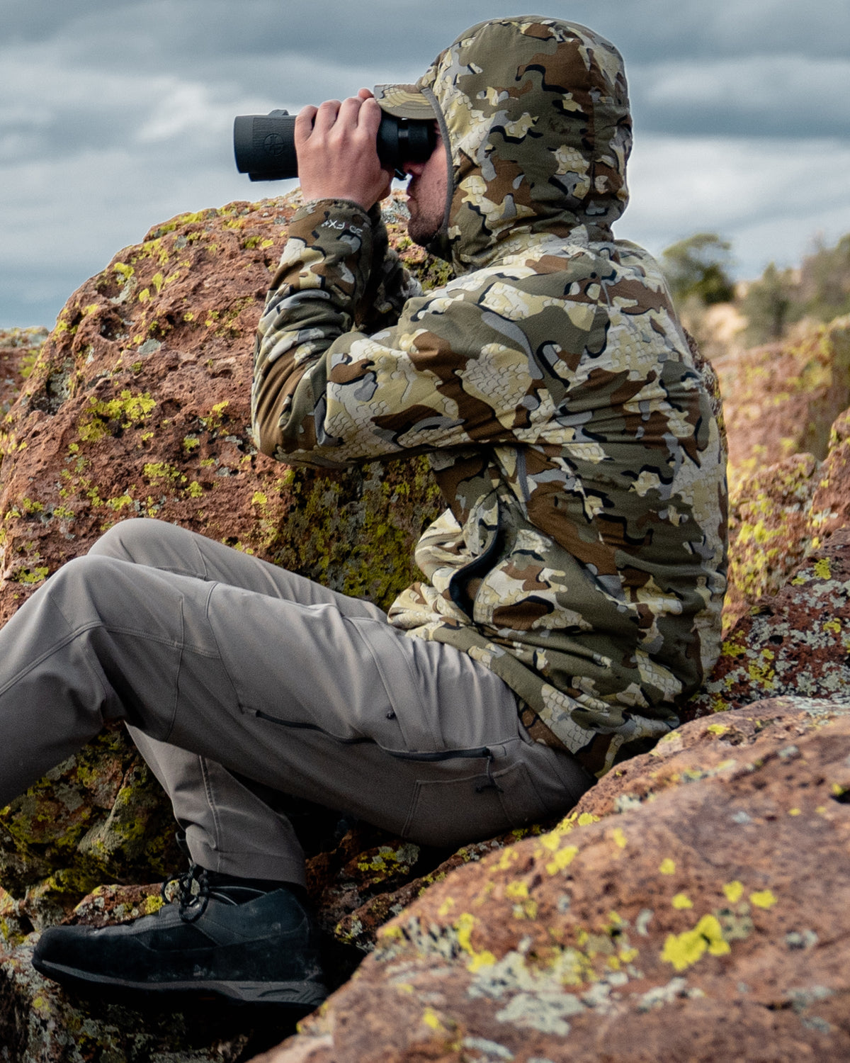 hunter sitting in rocks using binoculars wearing Kenai hooded Jacket in Valo Camouflage