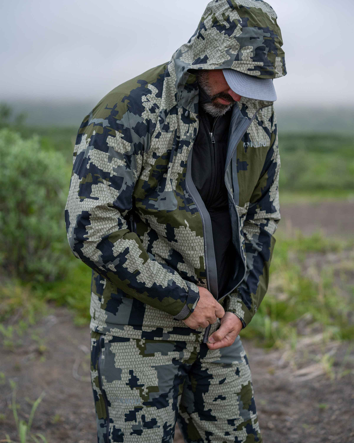 Hunter in field zipping up Chugach TR Rain Jacket in Verde Camouflage
