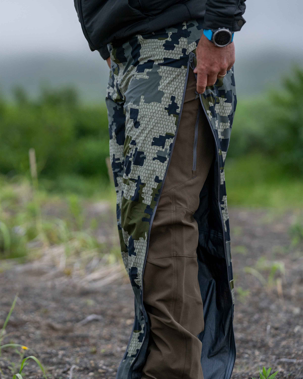 Hunter in field unzipping full-length leg zipper on Chugach TR Rain Pant in Verde Camouflage
