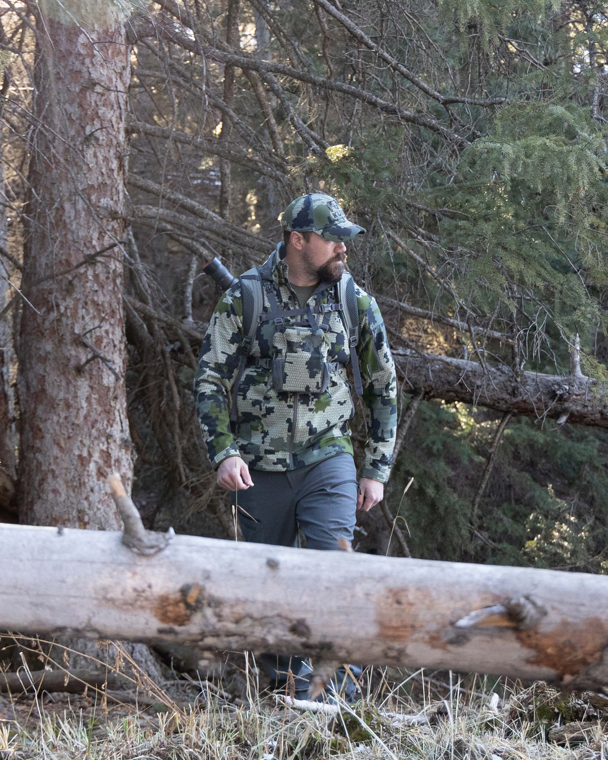 Hunter Walking Through Pine Trees Wearing Guide PRO Hooded Jacket in Verde Camouflage