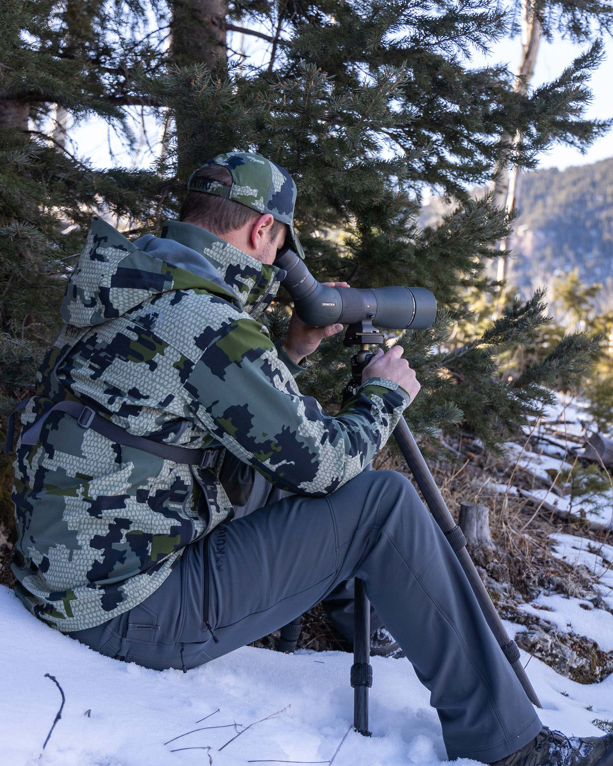 Hunter Sitting in Snow in Front of Pine Tree Wearing Guide PRO Hooded Jacket in Verde Camouflage While Watching Game Through Spotting Scope