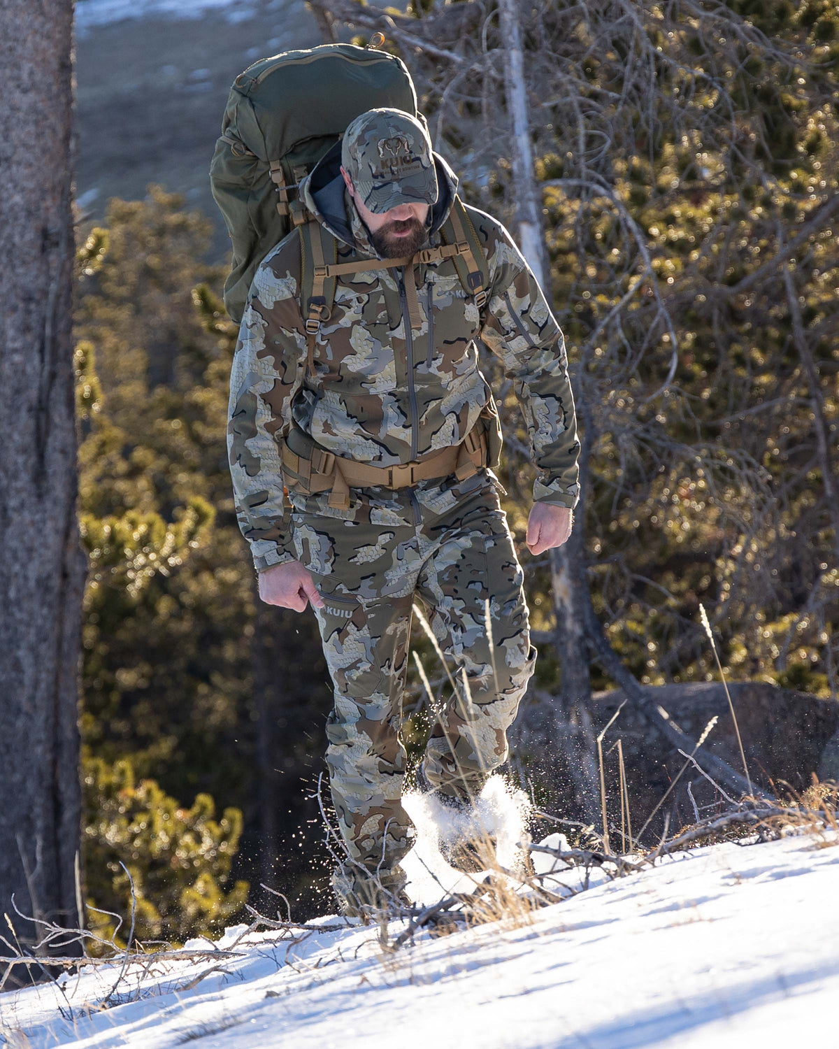 Hunter Walking Up Snowy Hillside in Wooded Terrain Wearing Guide PRO Hooded Jacket in Valo Camouflage