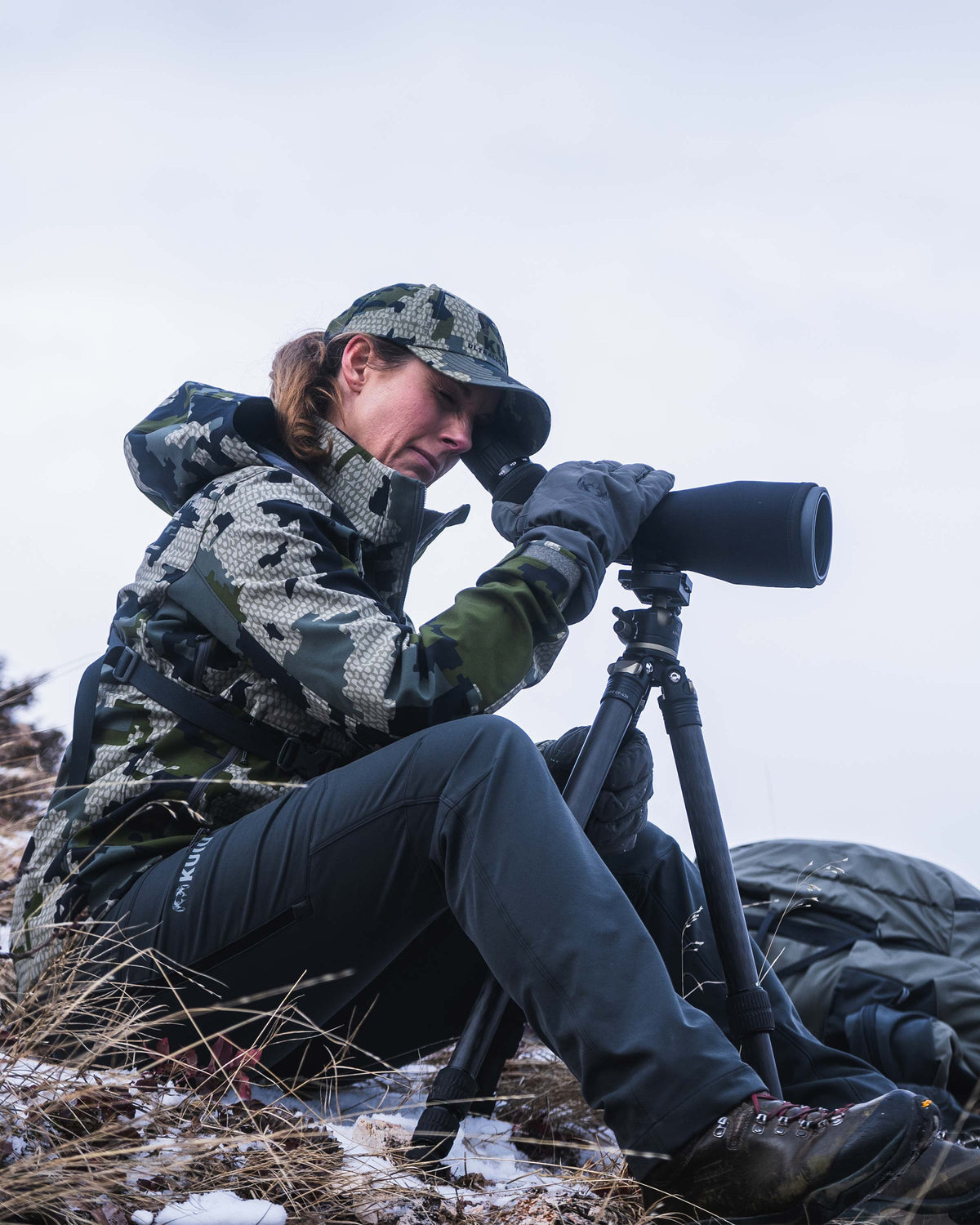 Side view of seated female hunter wearing Women's Guide PRO Hooded Jacket in Verde camouflage while looking through spotting scope