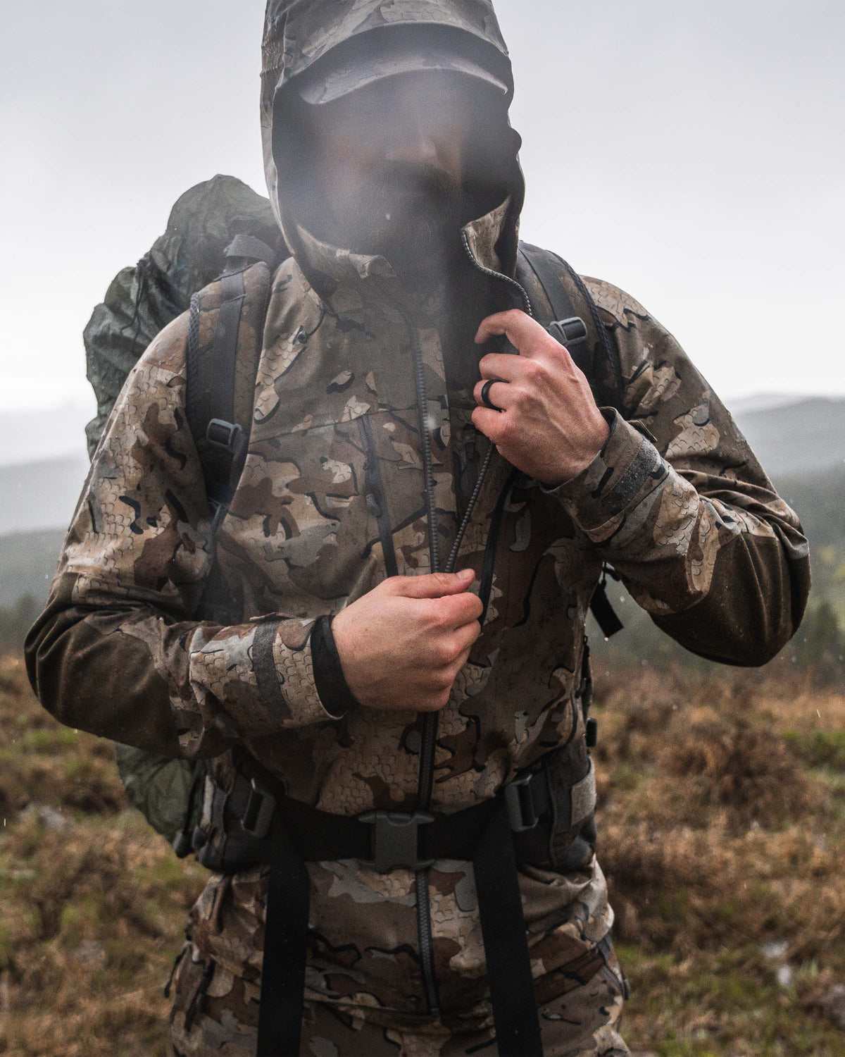 Frontal view of hunter wearing Yukon TR Rain Jacket in Valo Camouflage zipping up the jacket in the rain while wearing a pack