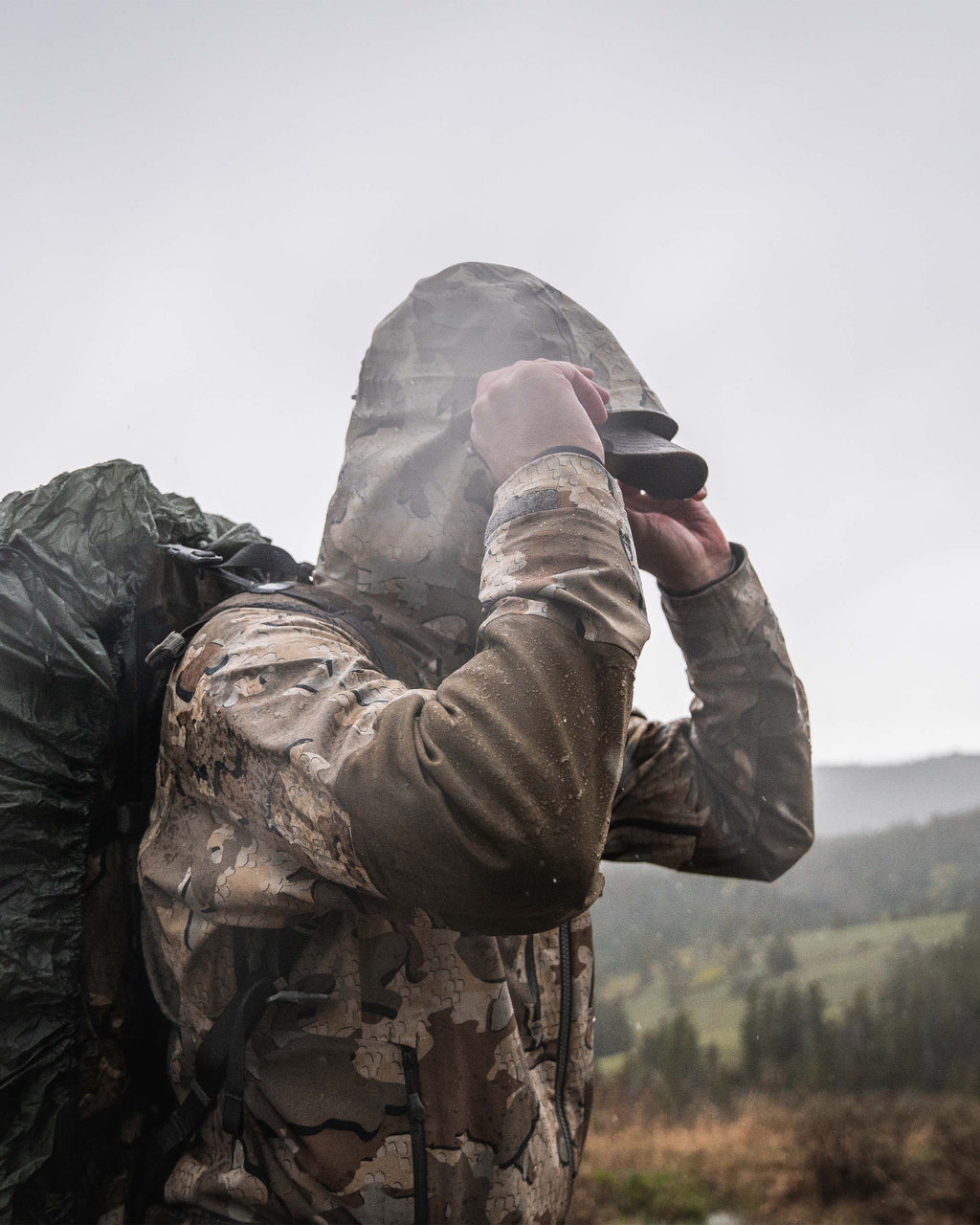 Side profile view of hunter wearing Yukon TR Rain Jacket in Valo camouflage adjusting hood visor while standing in the rain
