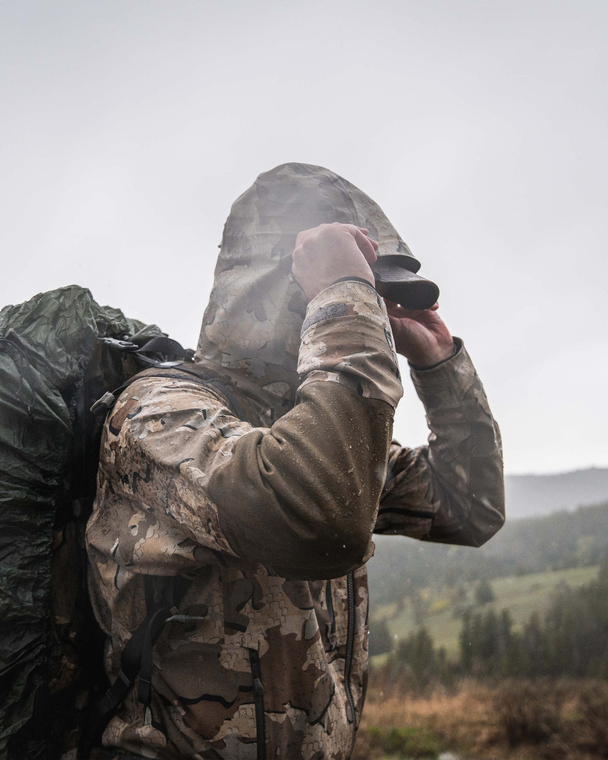Side profile view of hunter wearing Yukon TR Rain Jacket in Valo camouflage adjusting hood visor while standing in the rain