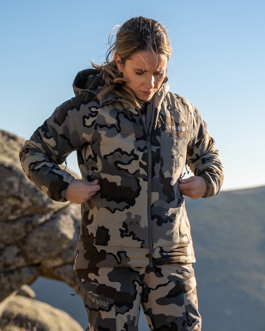 Female hunter unzipping both hand pockets on Women's Kenai Hooded Jacket in Vias Camouflage