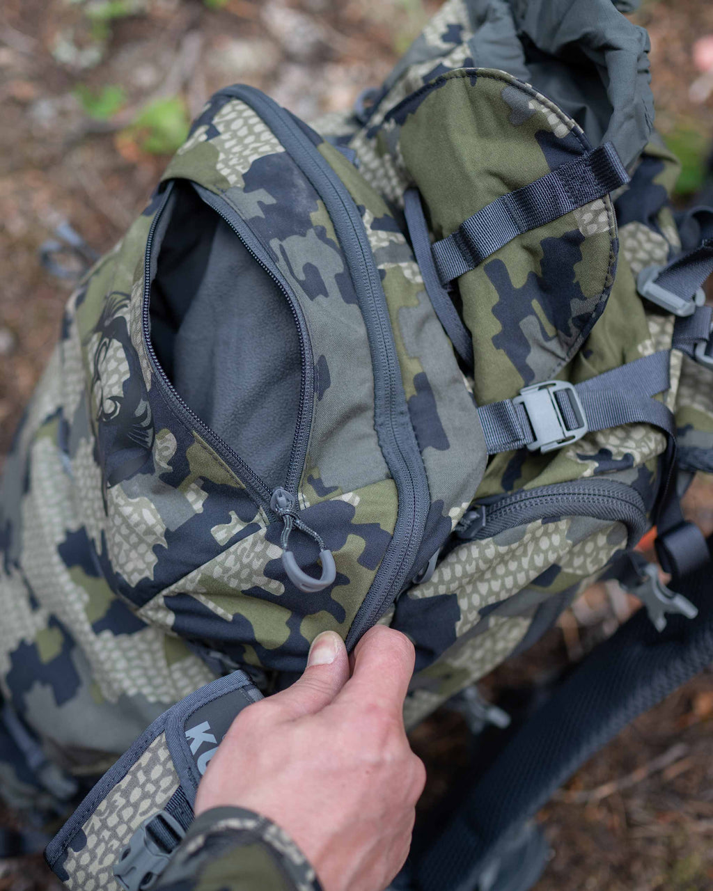 Aerial a hunter unzipping the front pocket on the Venture Divide 3000 Pack in Verde Camouflage