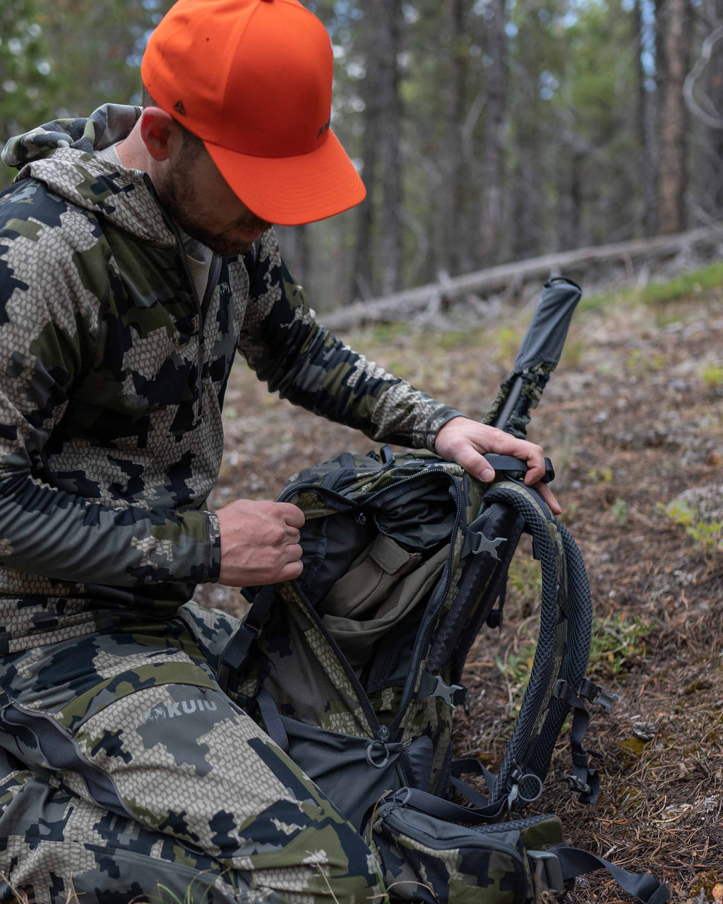 Hunter Kneeling in Wooded Area Opening Main Compartment on Venture Divide 3000 in Verde Camouflage