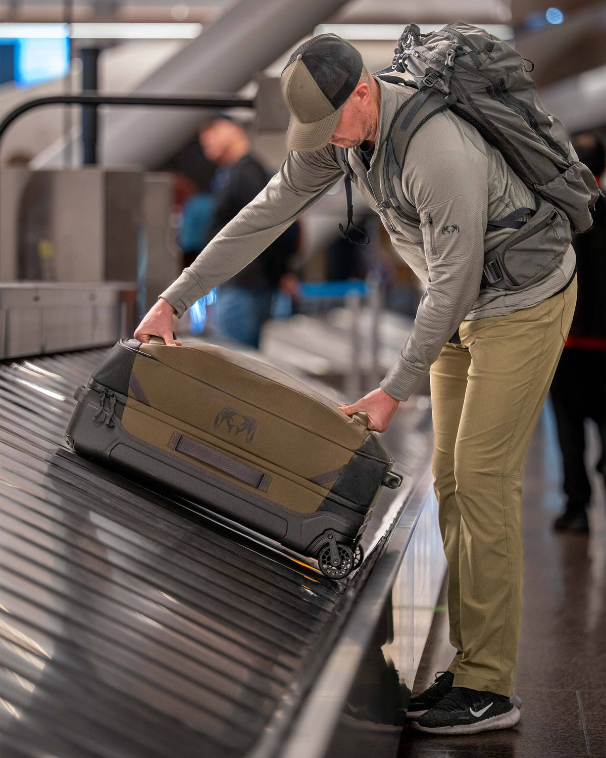 Man standing beside a luggage conveyor belt leaning forward and using both hands on the front handles to lift Waypoint 4500 Roller in Bourbon Brown.