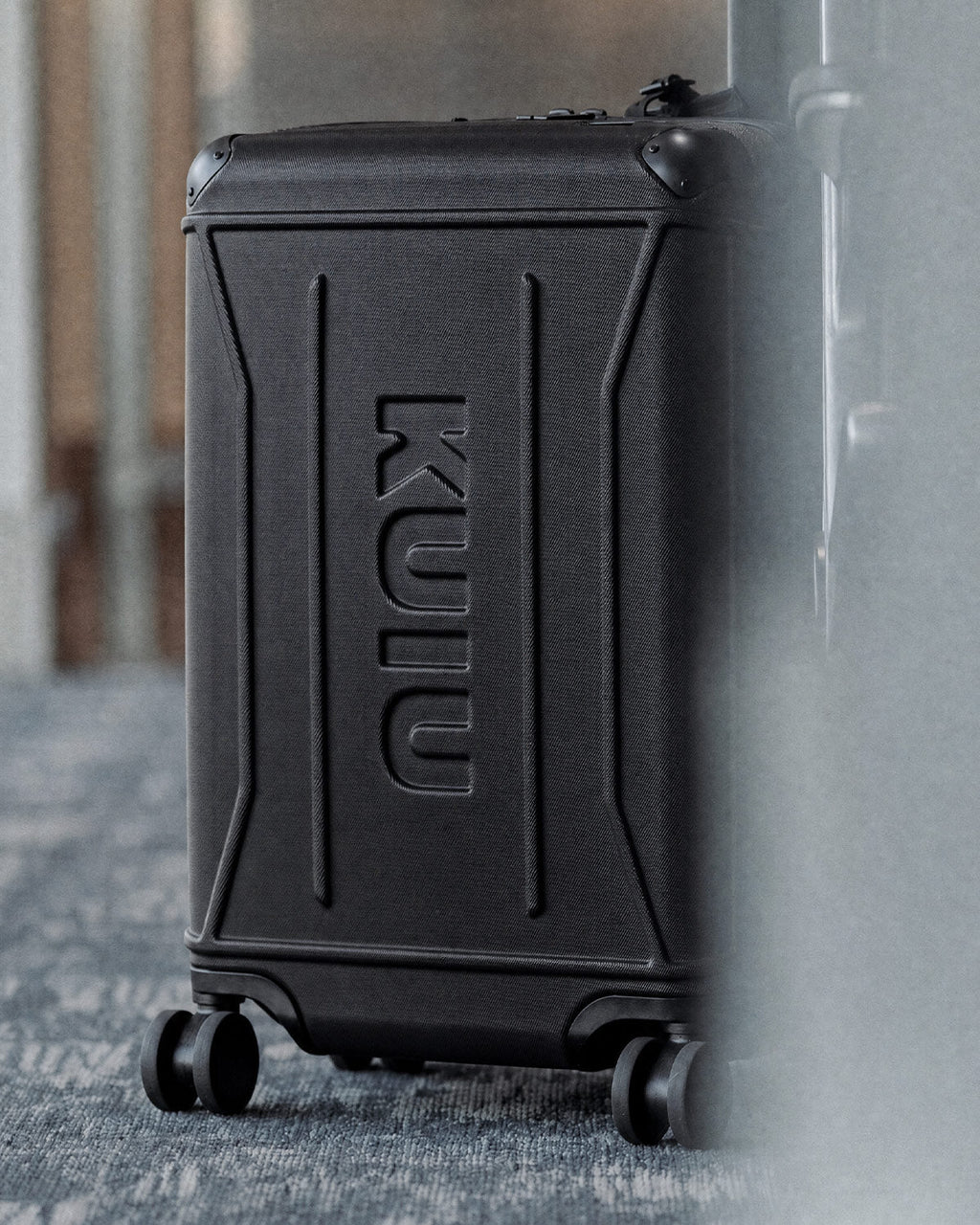 A black Hardline Spinner Carry-On in Black with a large embossed vertical KUIU logo on the front, photographed indoors on a textured gray floor with a blurred background.