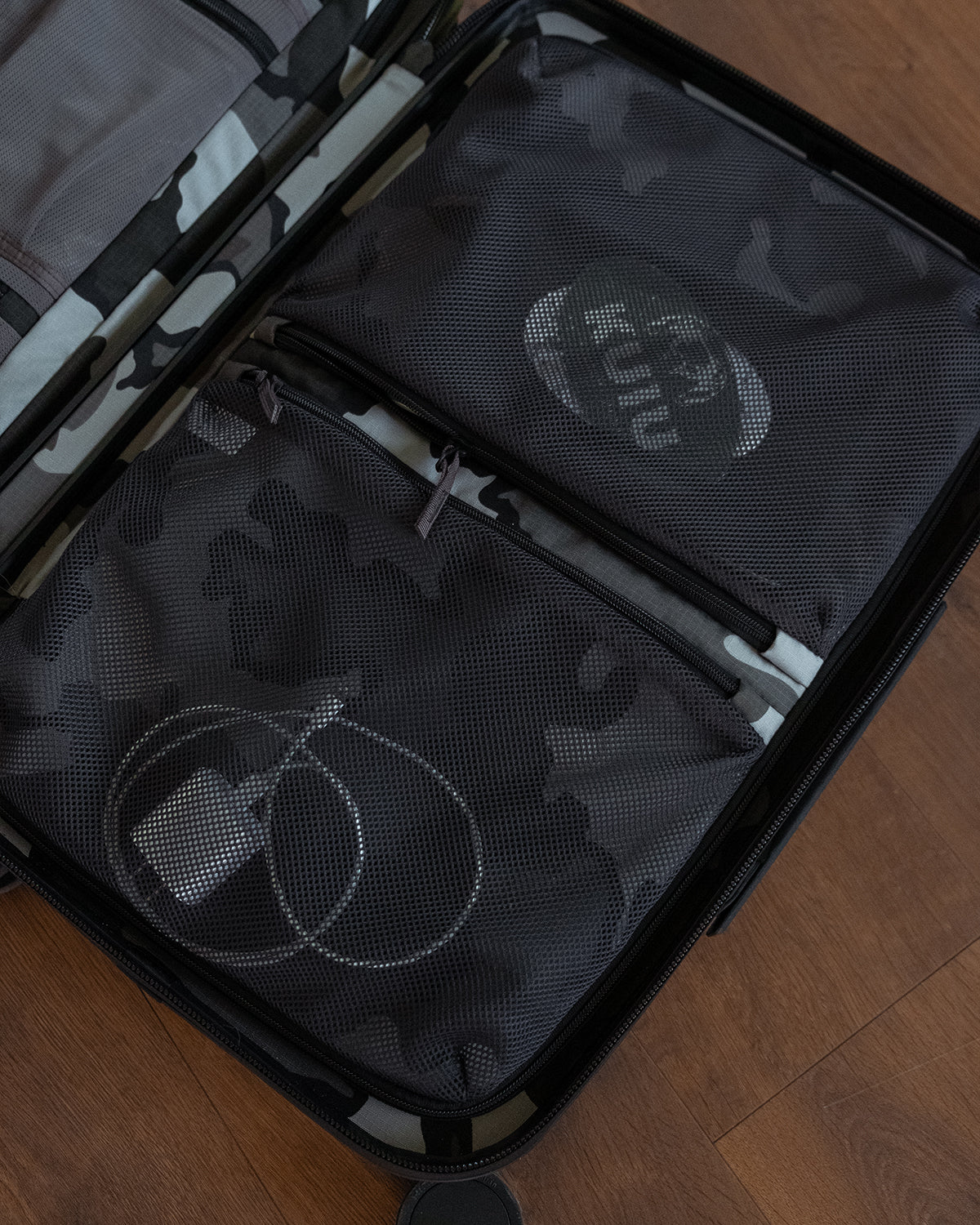 A close-up of an open Hardline Spinner Carry-On showing two dark gray camouflage mesh organizer pockets inside, with one pocket printed with the word 'KUIU' and the other holding a white cable. The suitcase is placed on a wooden floor.