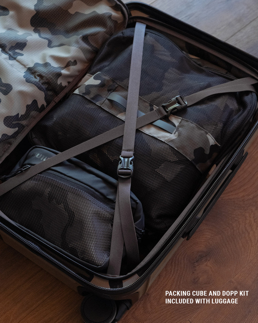 Vias camouflage packing cube and a Dopp kit inside an open Hardline Spinner Carry-On on a wooden floor, secured by a gray compression strap.