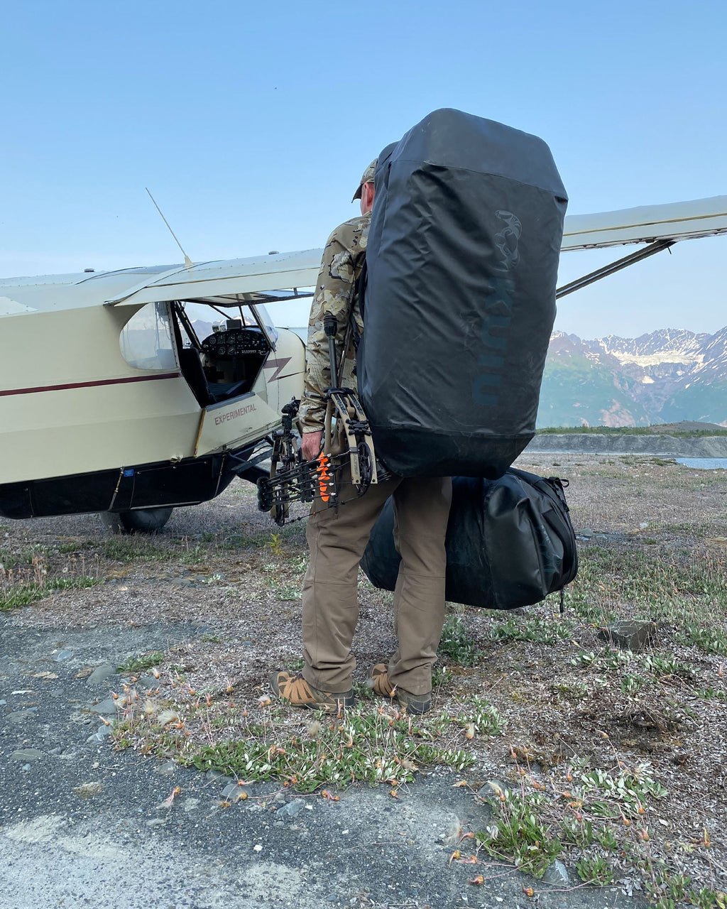 Quartering away view from the rear of a hunter wearing black Waypoint 10400 Duffel bag on back as he waits to board a small bush plane
